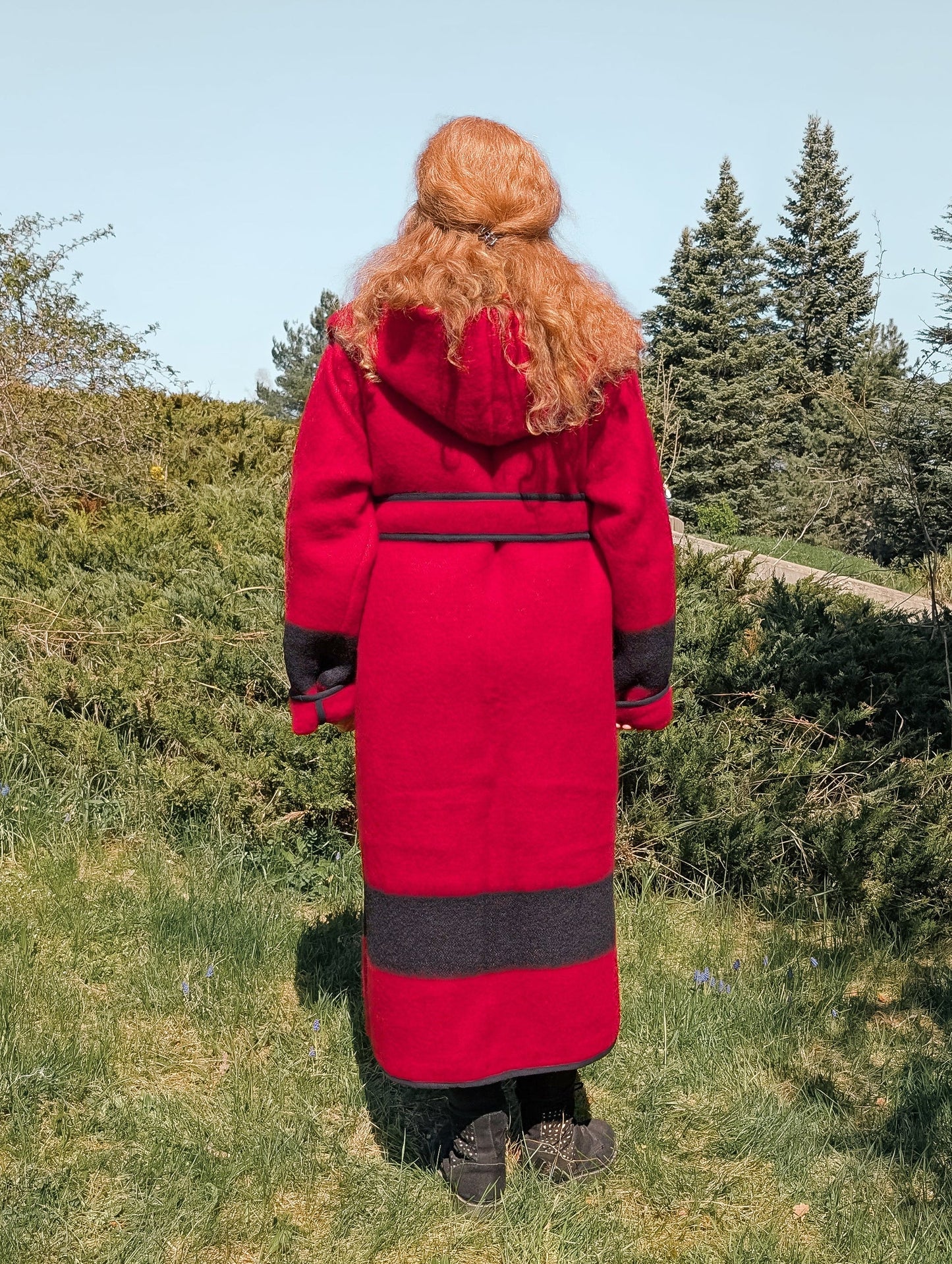 A woman posing backwards with a custom made long wool jacket in red with black stripes, hood and belt.