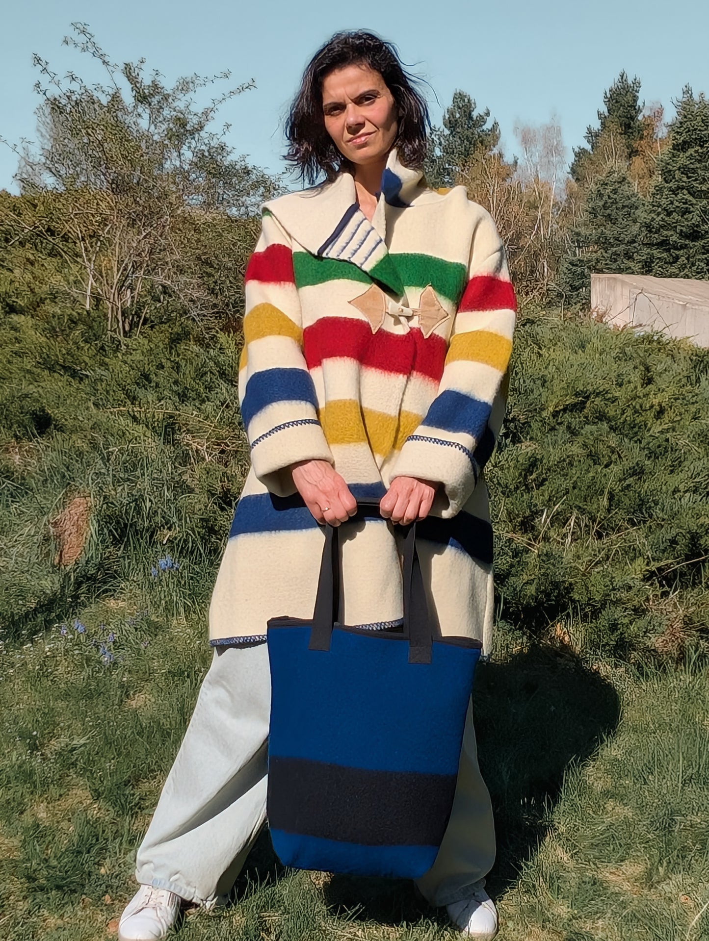Woman wearing a midi striped coat with a hood, and holding a matching bag, both upcycled from woolen blanket.
