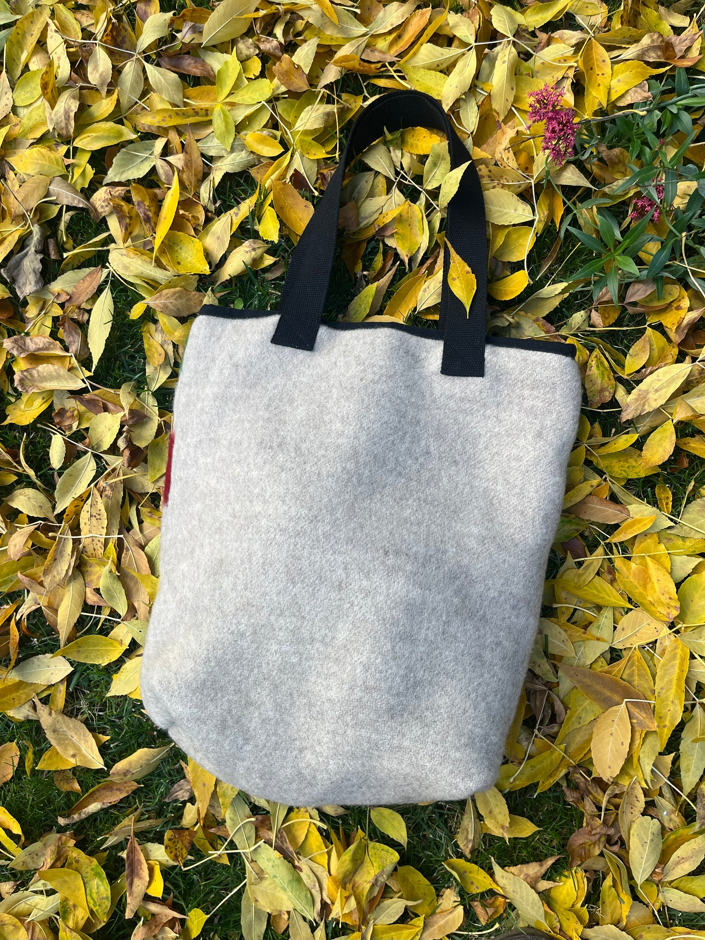 The natural gray backside of a handmade tote bag with black handles, laying on a bed of yellow autumn leaves