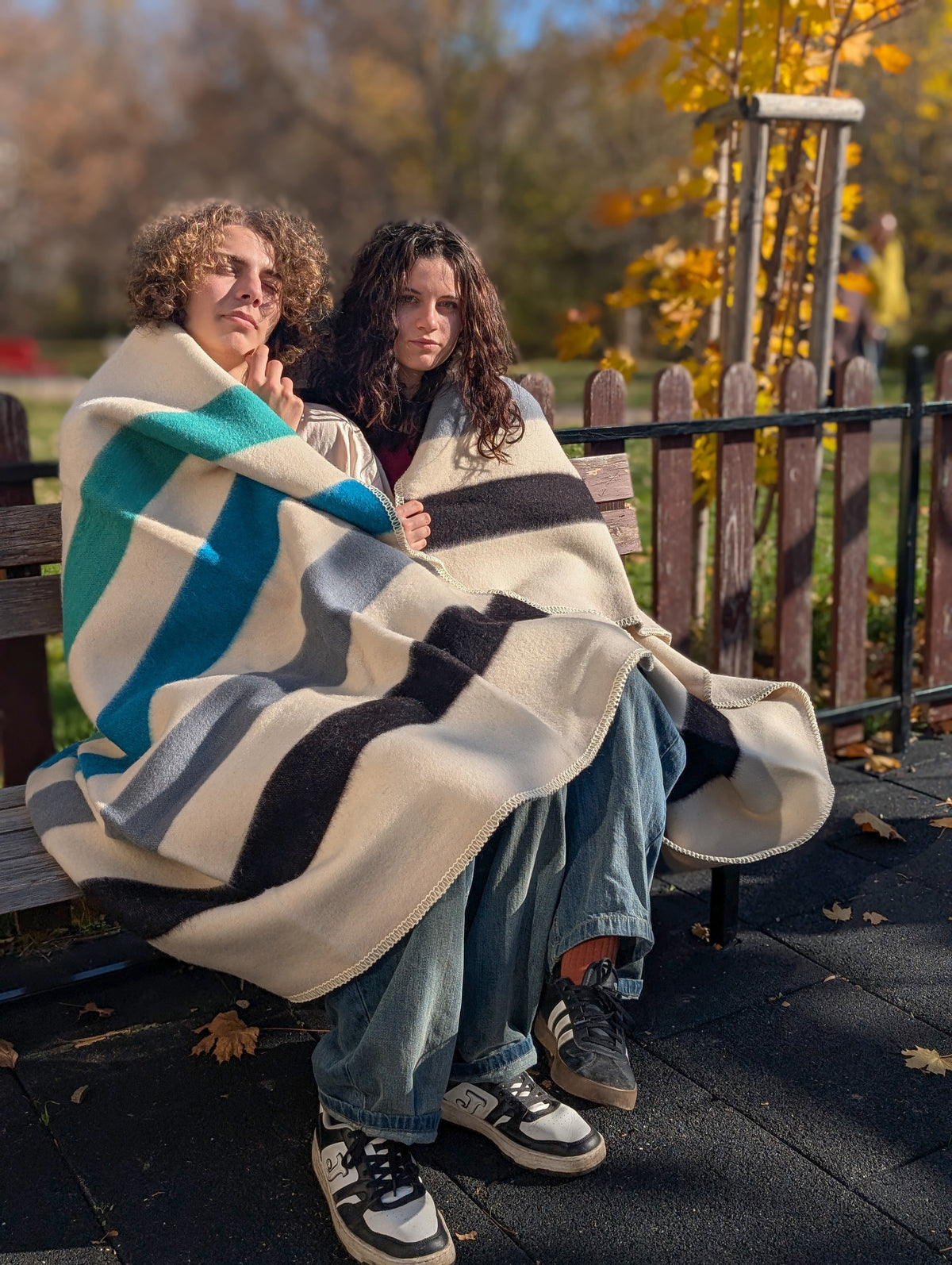 Two people sitting on a bench wrapped in an authentic handwoven multistriped blanket with autumn trees in the background.