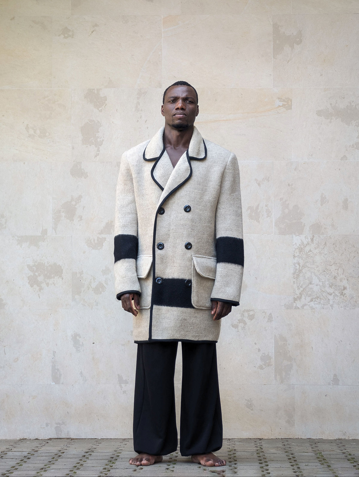 Front full-length shot of a model wearing a gray double-breasted wool peacoat with black pipe edging and black stripe accent.
