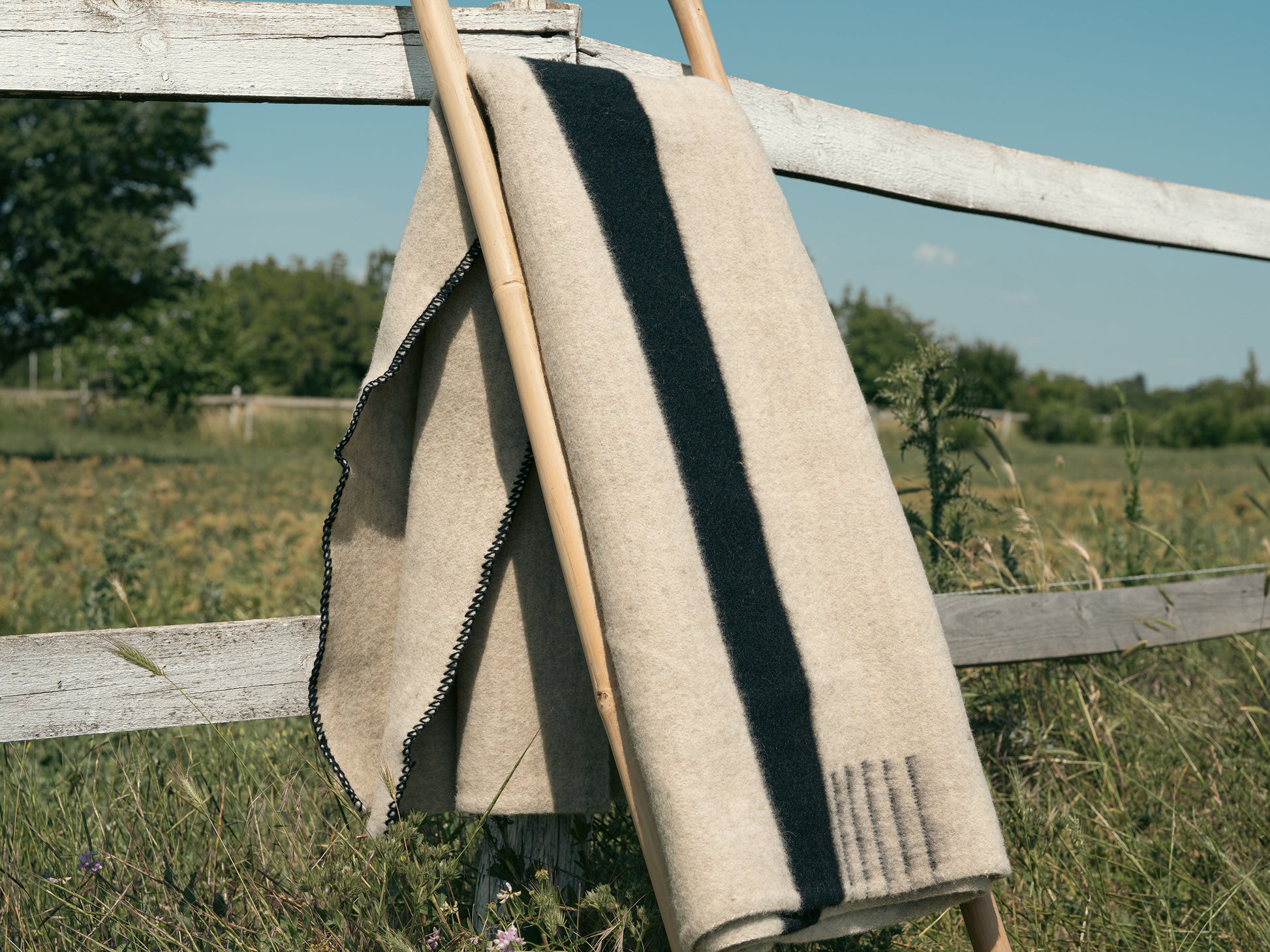 Light natural handwoven wool blanket with black stripes, folded near a farm fence.