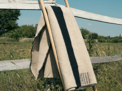 Light natural handwoven wool blanket with black stripes, folded near a farm fence.