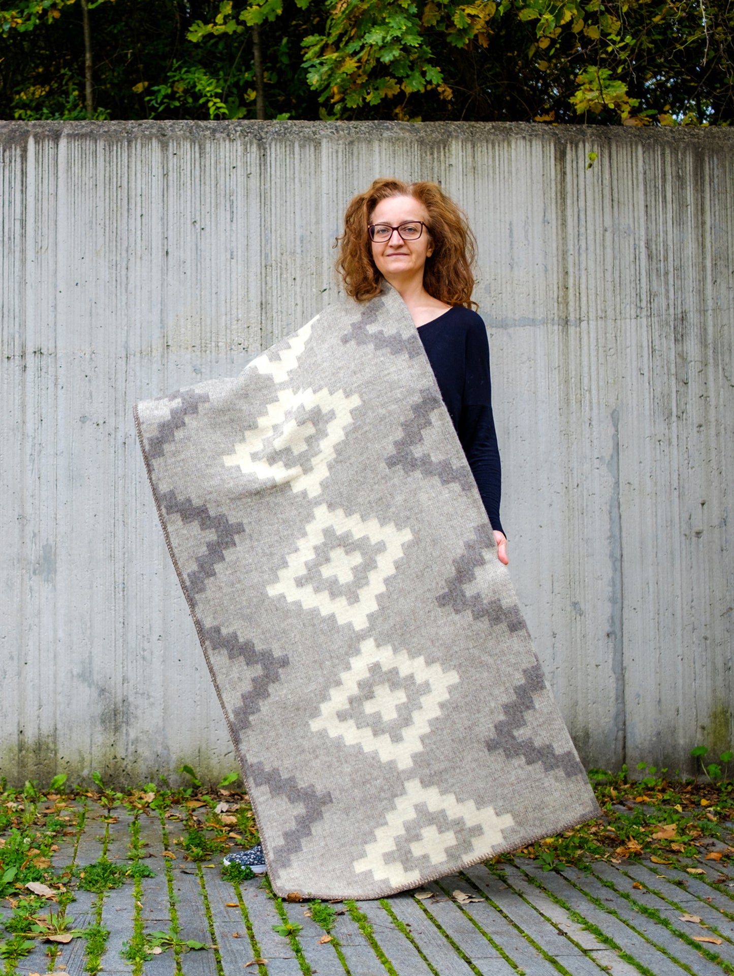Woman holding a large heavyweight undyed wool blanket in soft gray natural colors with stepped geometric pattern.