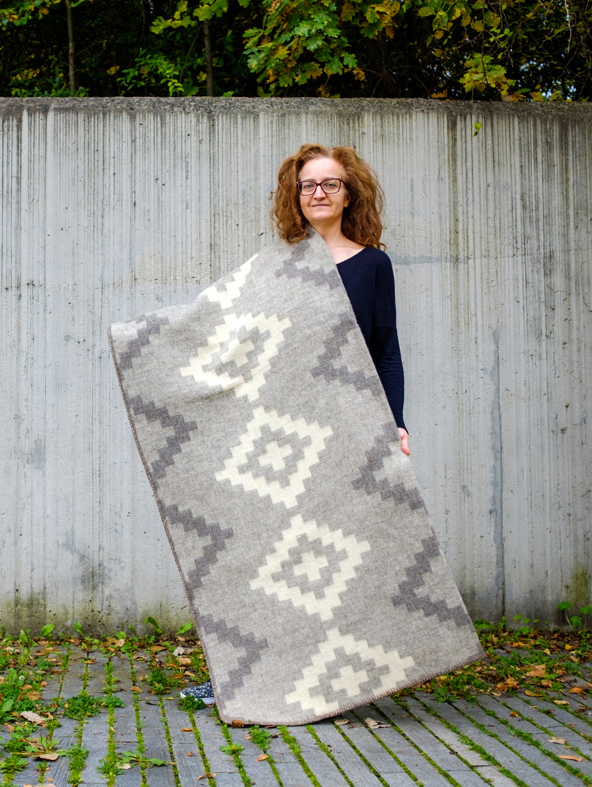 Woman holding a large heavyweight undyed wool blanket in soft gray natural colors with stepped geometric pattern.