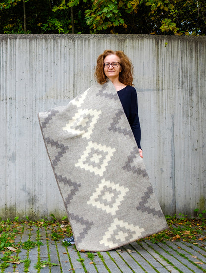 Woman holding a large heavyweight undyed wool blanket in soft gray natural colors with stepped geometric pattern.