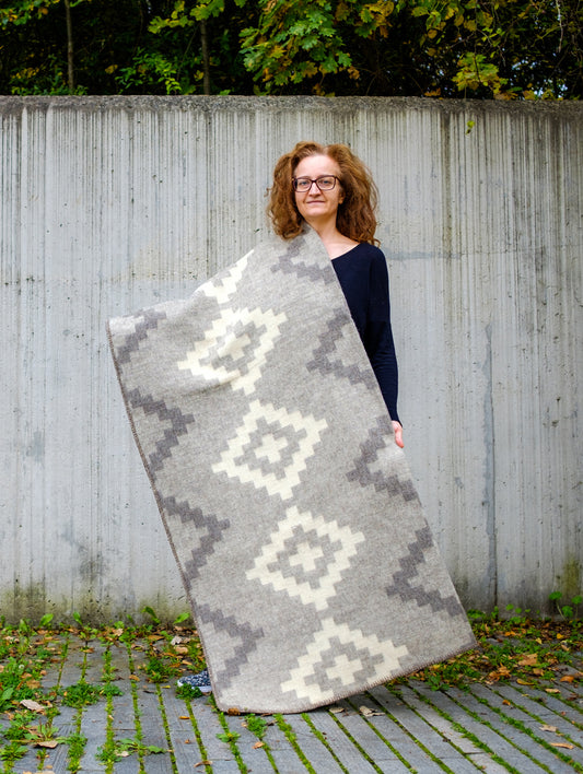 Woman holding a large heavyweight undyed wool blanket in soft gray natural colors with stepped geometric pattern.