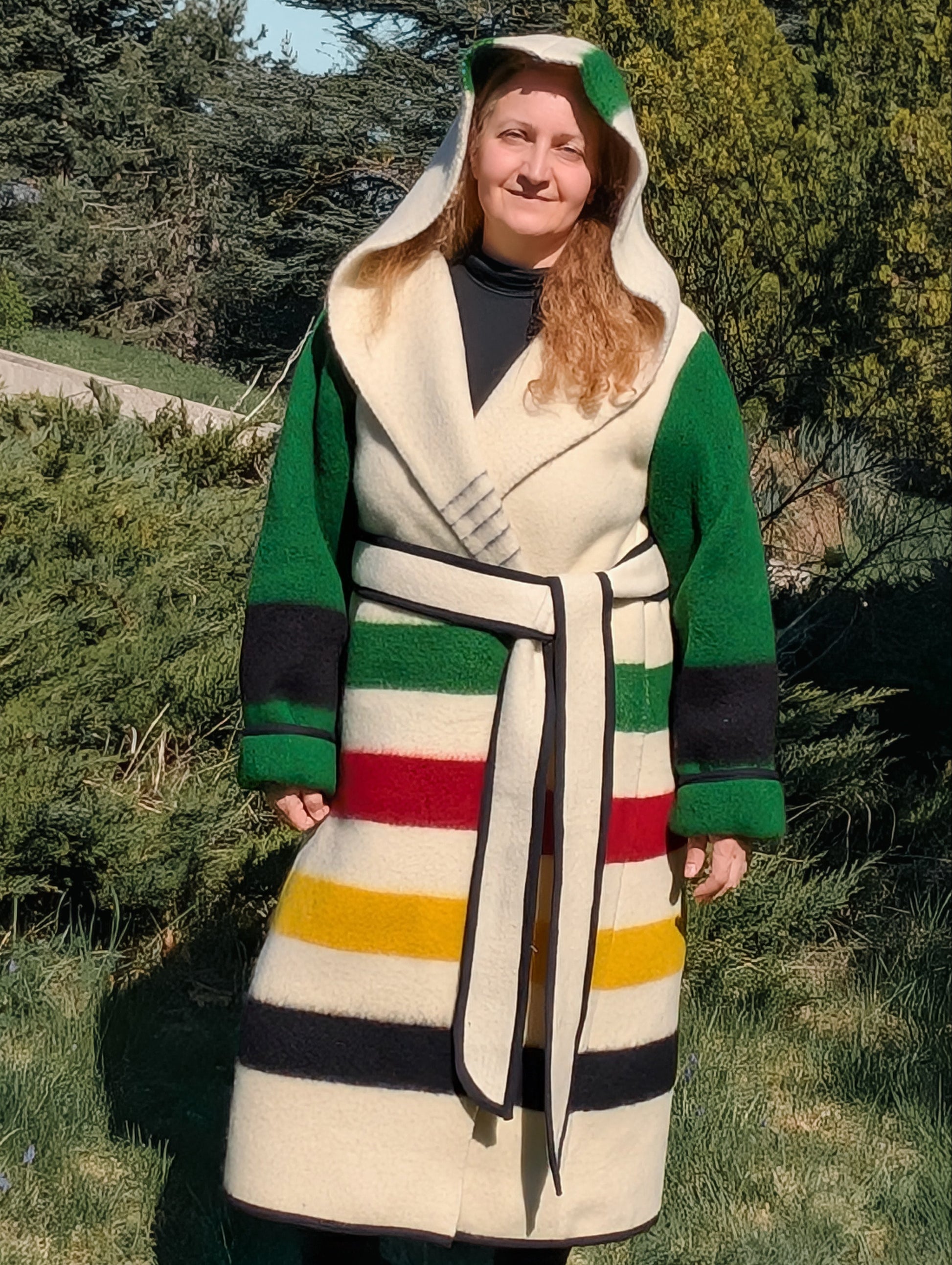Woman wearing a long hooded capote coat with colorful stripes and green sleeves, upcycled from a single woolen blanket.