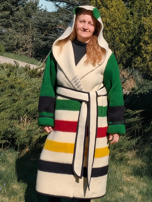 Woman wearing a long hooded capote coat with colorful stripes and green sleeves, upcycled from a single woolen blanket.