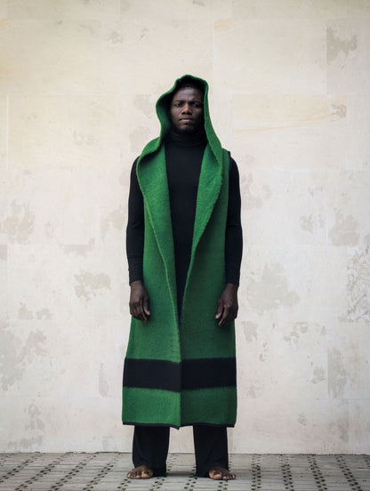 A man wearing a long green hooded wrap coat with black stripes over a black outfit.