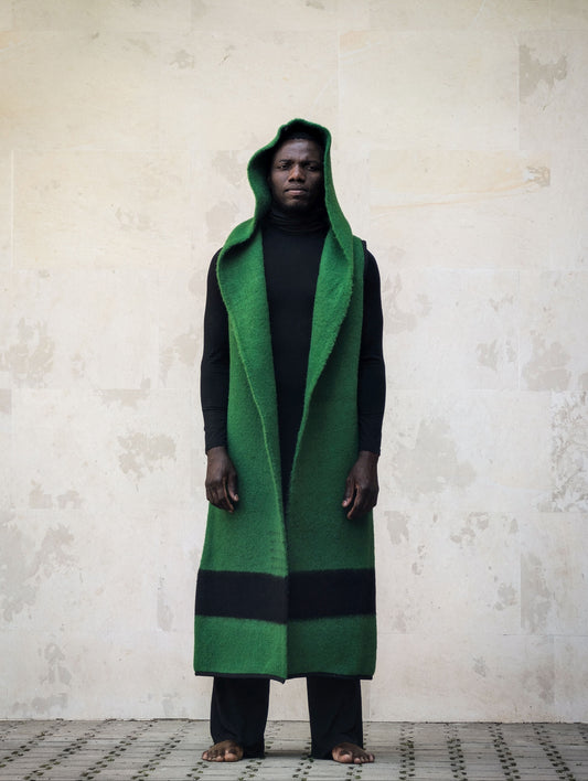 A man wearing a long green hooded wrap coat with black stripes over a black outfit.