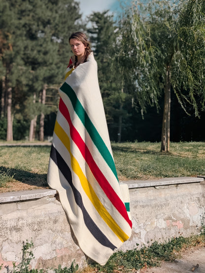 A standing woman wrapped in a cozy warm off white wool blanket with multicolor stripes.