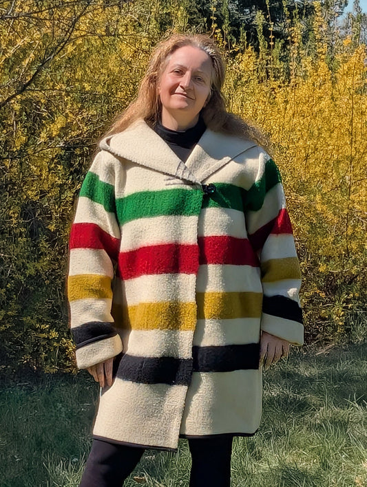 A woman wearing a midi swing capote coat upcycled from a multistripe blanket. The model features toggle closure and a wide shawl hoodie. 