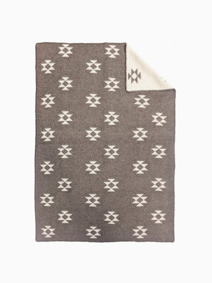 A handmade woven throw blanket with a repeating white geometric star pattern on a taupe-brown background, featuring a folded corner that reveals a cream-colored reverse side