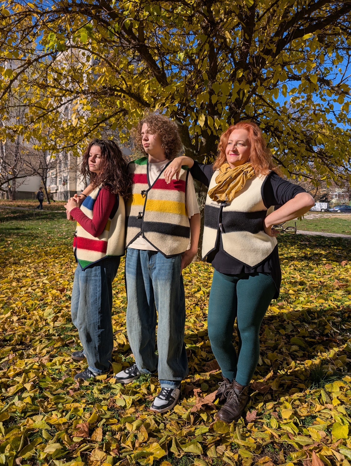 Three people standing together in a park with autumn leaves, and all three wearing similar stylish vests made from various upcycled wool blankets.