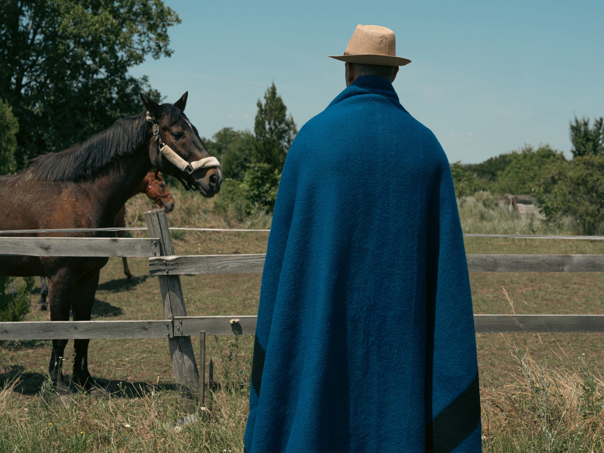Man wrapped in a blue organic wool blanket, with a hat, standing next to a horse in a field