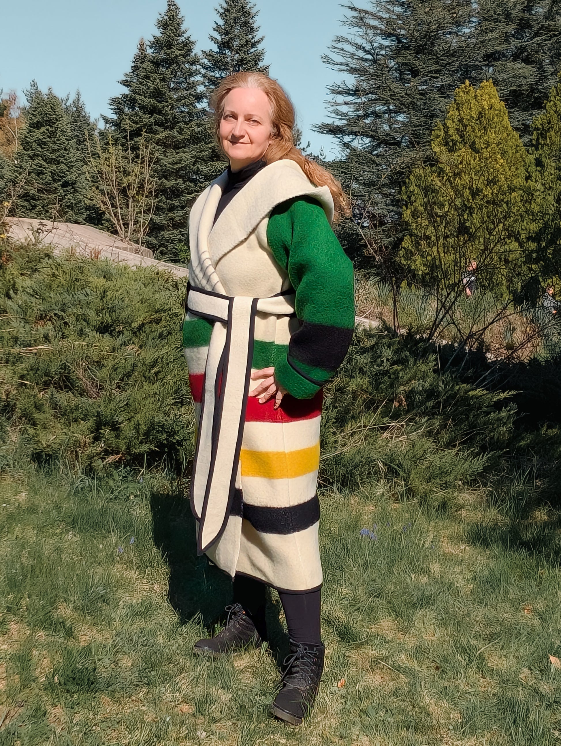 Woman wearing a long hooded blanket jacket with a belt, with multi stripe pattern and green sleeves.