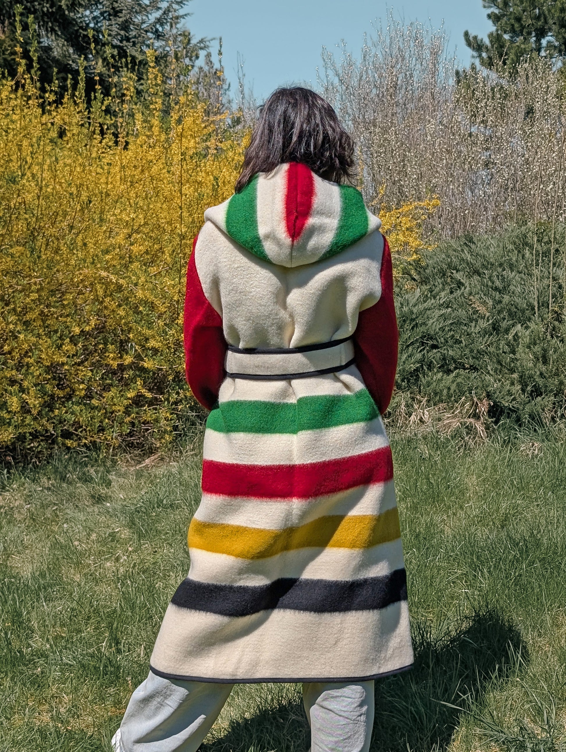 Woman wearing a wrap hooded coat, cinched with a belt, featuring iconic colorful stripes. The model is m,ade from repurposed single wool blanket.