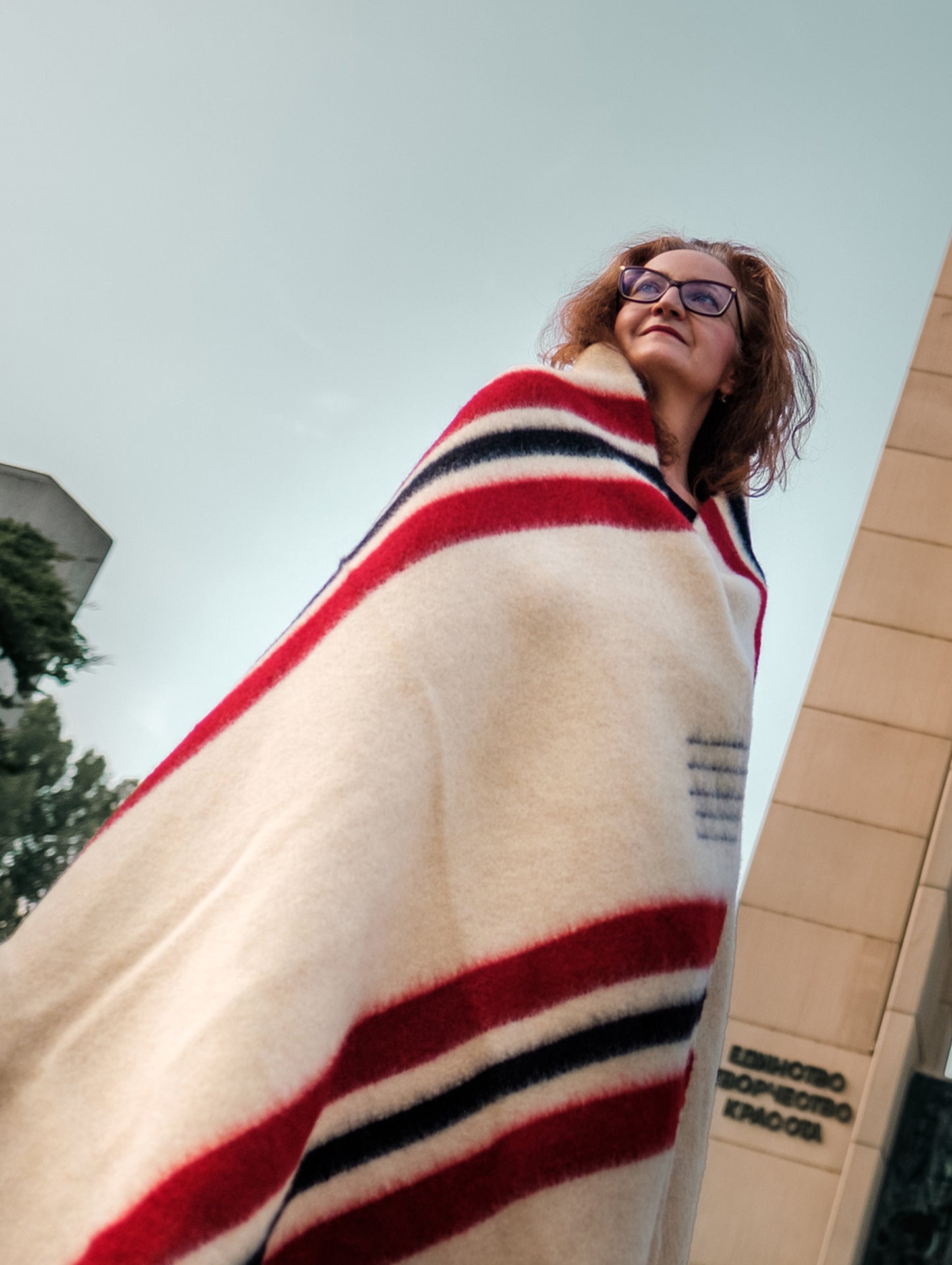 A ginger woman wrapped in a wool blanket featuring red and black stripes, posing in out in a park with a monument in the background