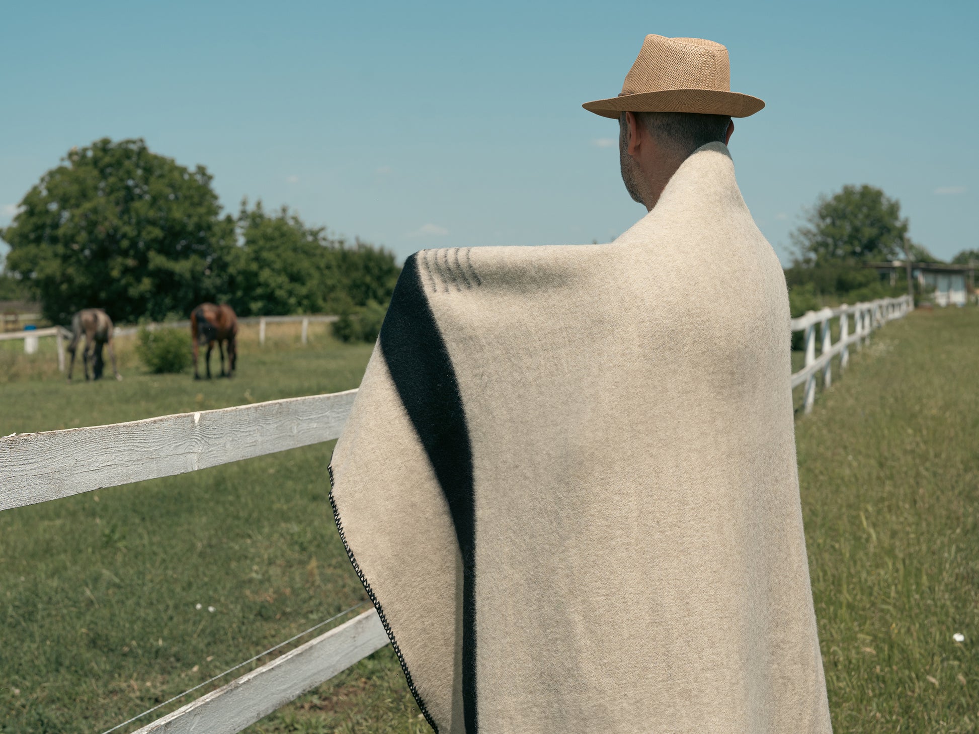A man with a hat posing wrapped with a beautiful camp blanket out near a ranch.
