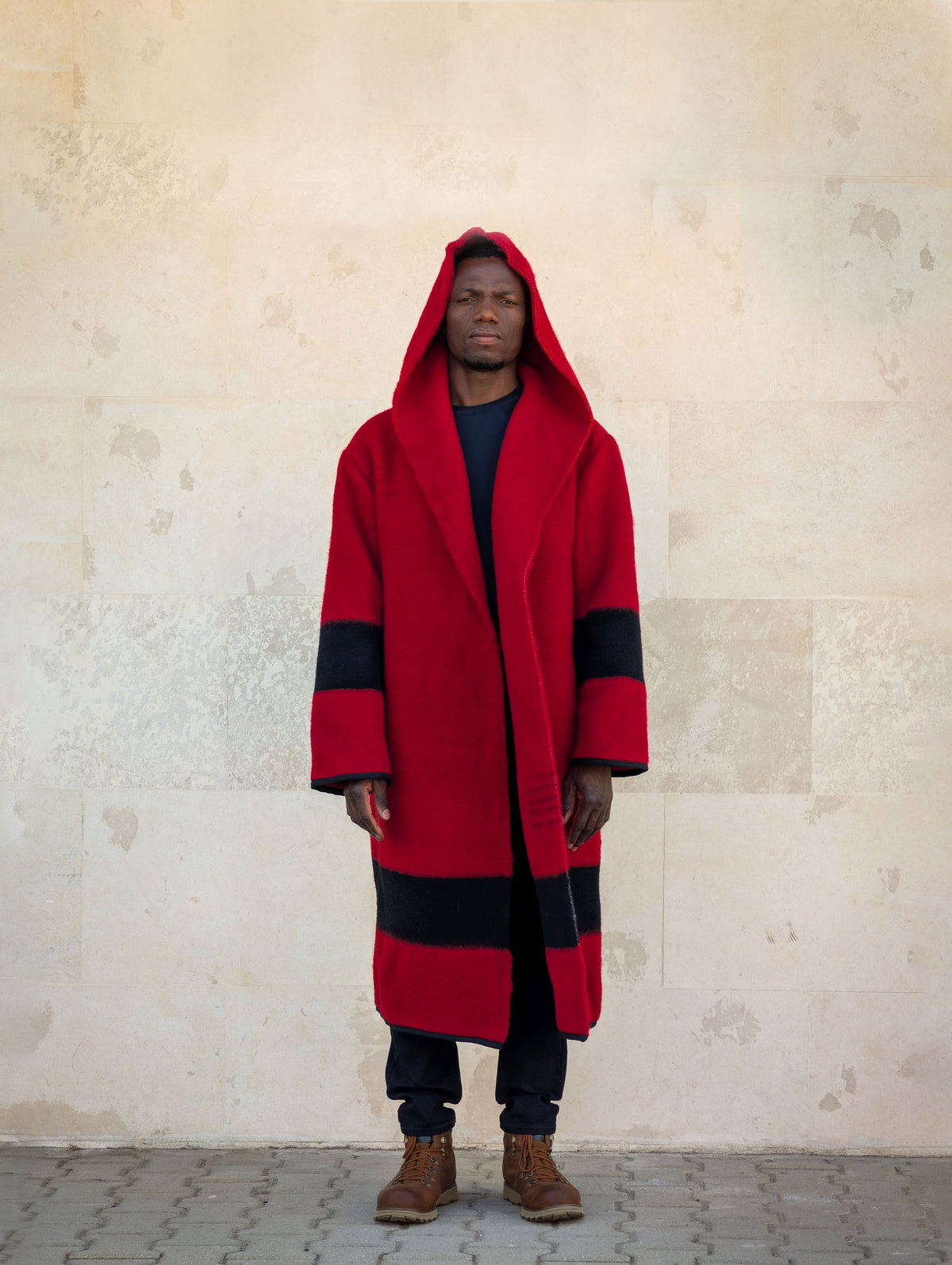 Model wearing a long unisex hooded capote coat tailored from handwoven wool blanket in solid red with a black stripe.