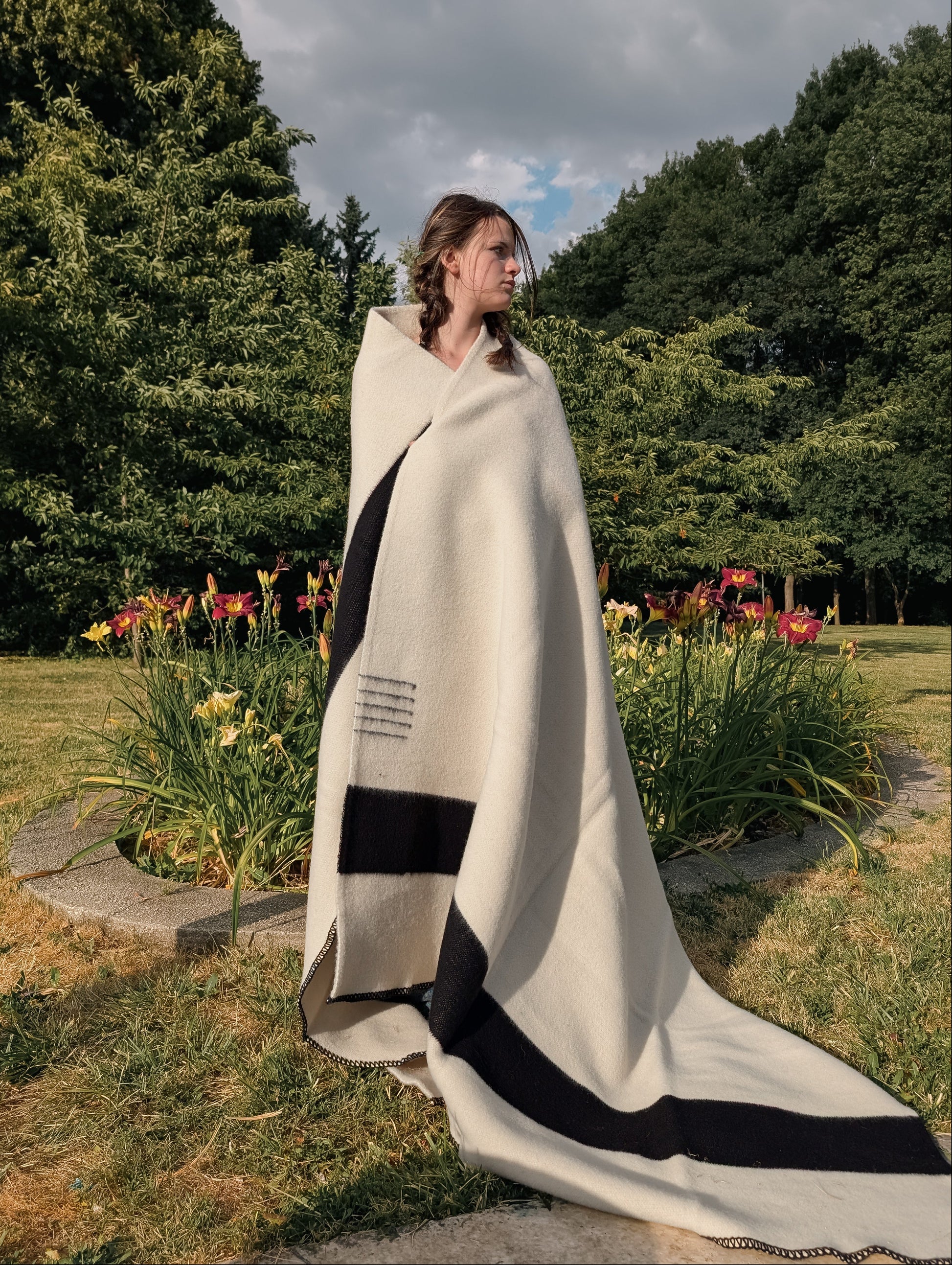Standing girl wrapped in a handwoven cream woolen blanket with black stripes in park setting