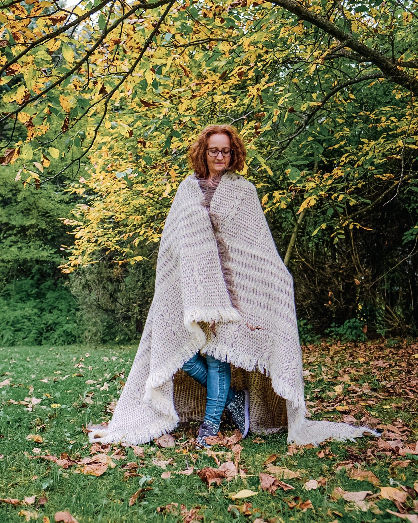 Woman wrapped in a luxurious undyed structured plaid wool throw in natural earth tones, finished wit fringes, standing out in the wild forest.