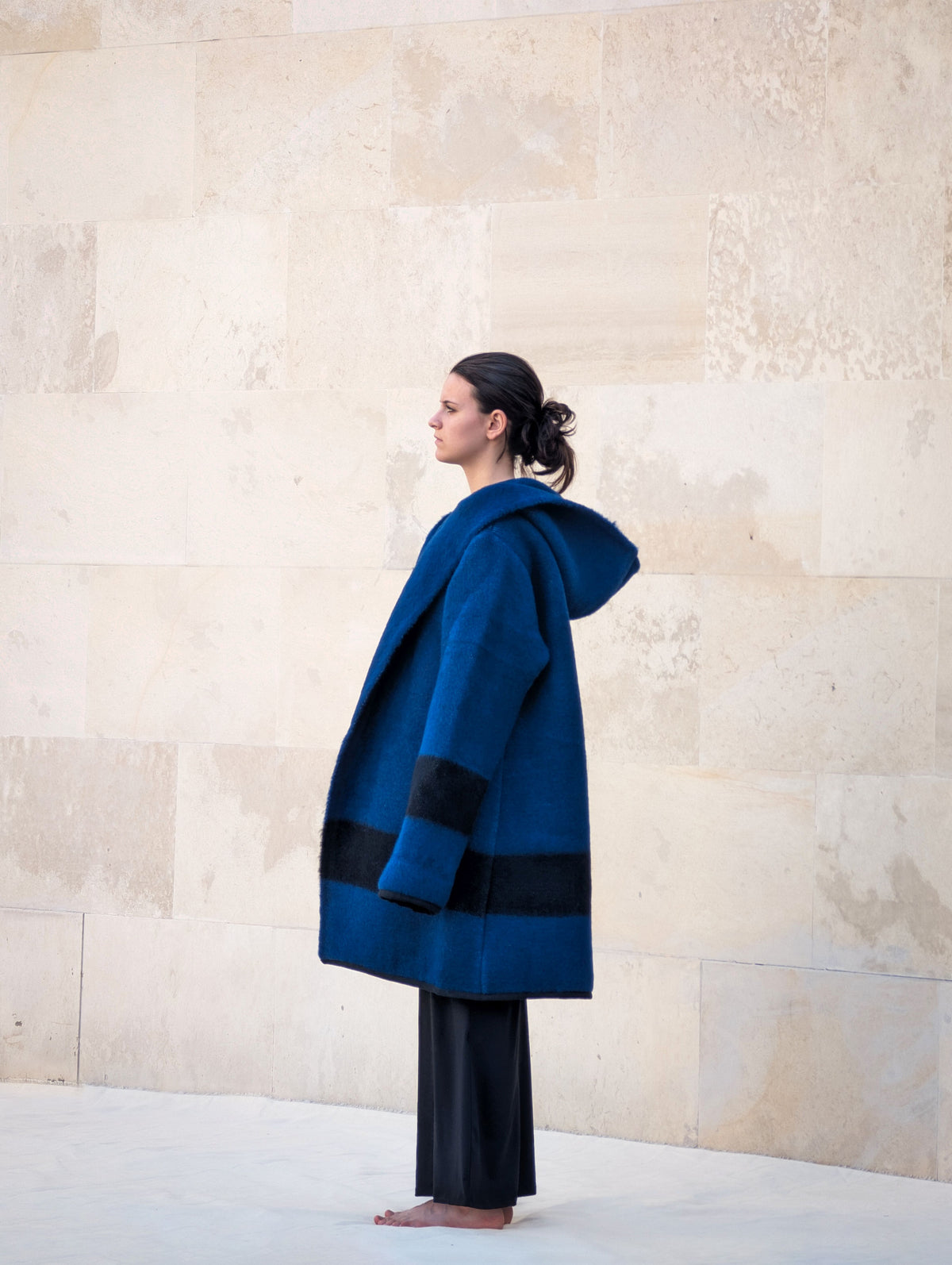 Side profile of a woman wearing an oversized blue wool capote coat with black stripe, hood raised.