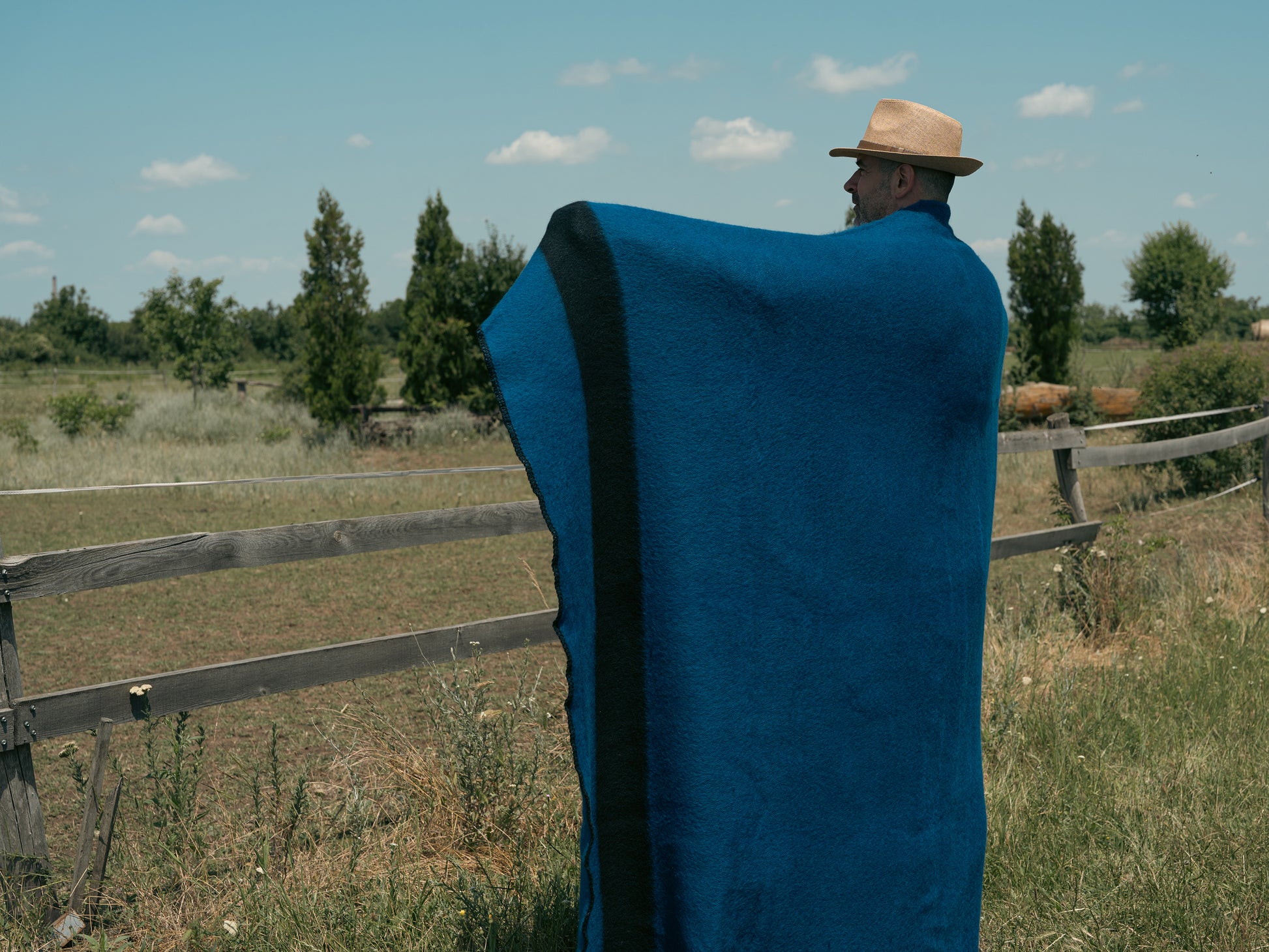Person wrapped in a blue stripe woven blanket standing in a field with a wooden fence and trees in the background.