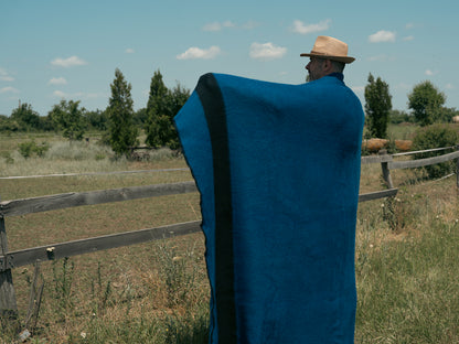 Person wrapped in a blue stripe woven blanket standing in a field with a wooden fence and trees in the background.