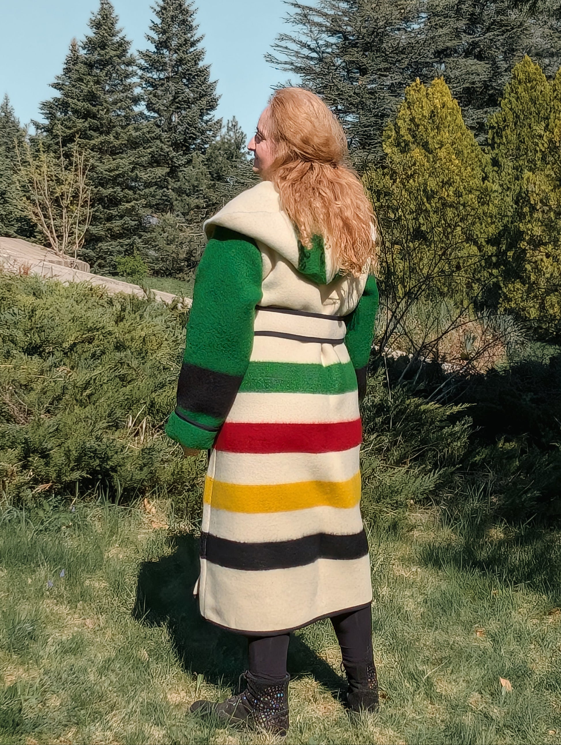 Woman posing backwards with a hooded blanket overcoat with colorful stripes and green sleeves. 