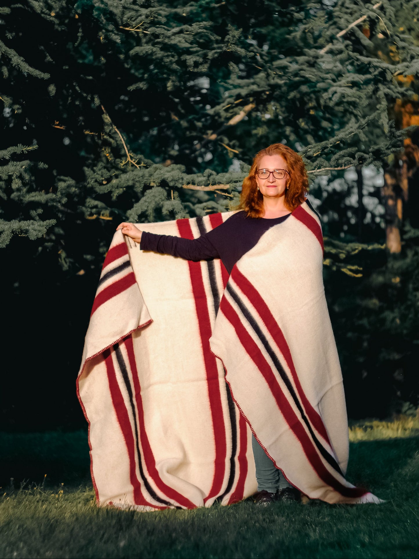 Woman wrapped in an off white wool striped Blanket with red and black stripes, and standing in front of some pine trees in a park