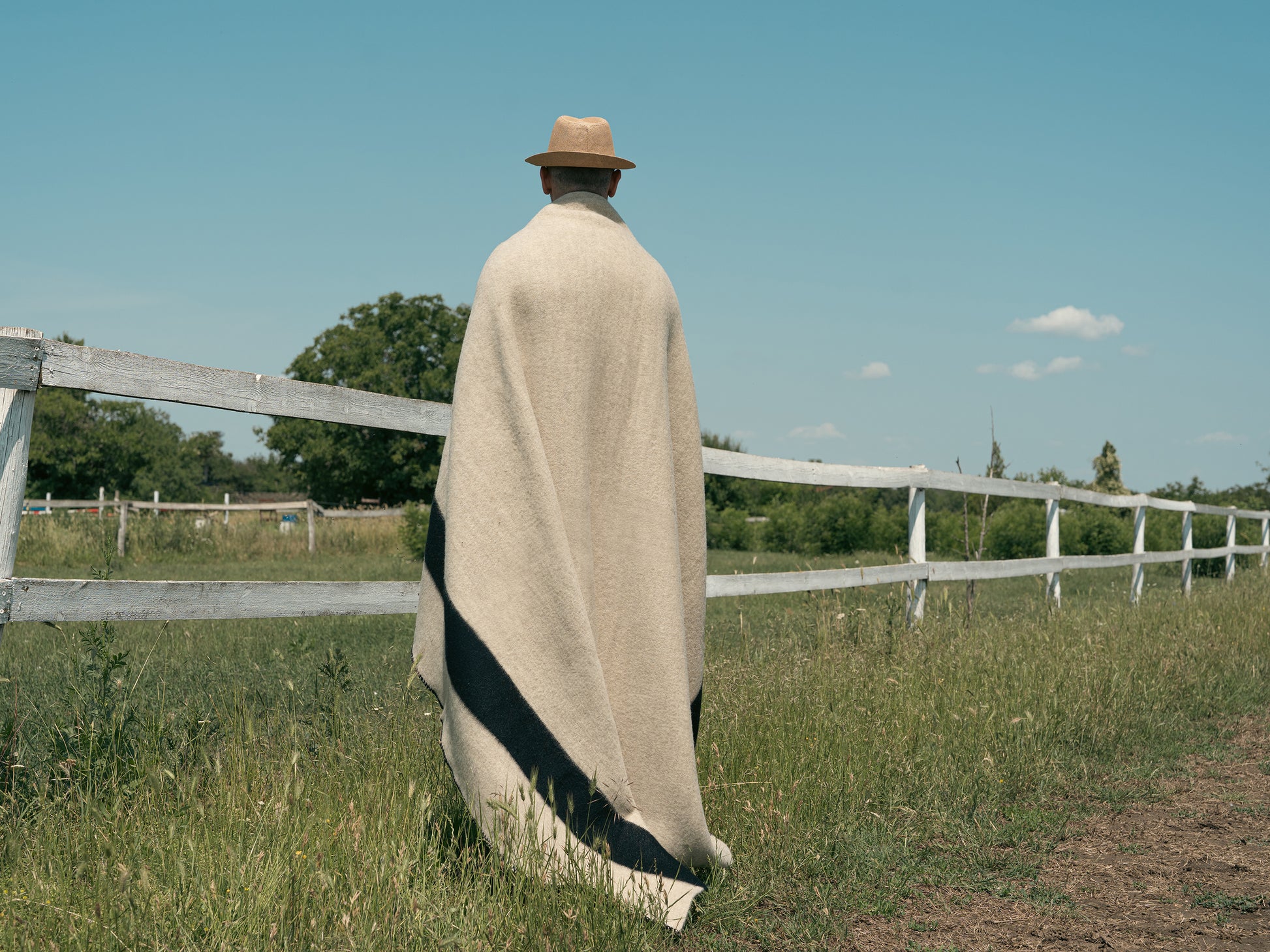 A man standing out near a fence, wrapped in a native american blanket handwoven from pure eco sustainable wool.