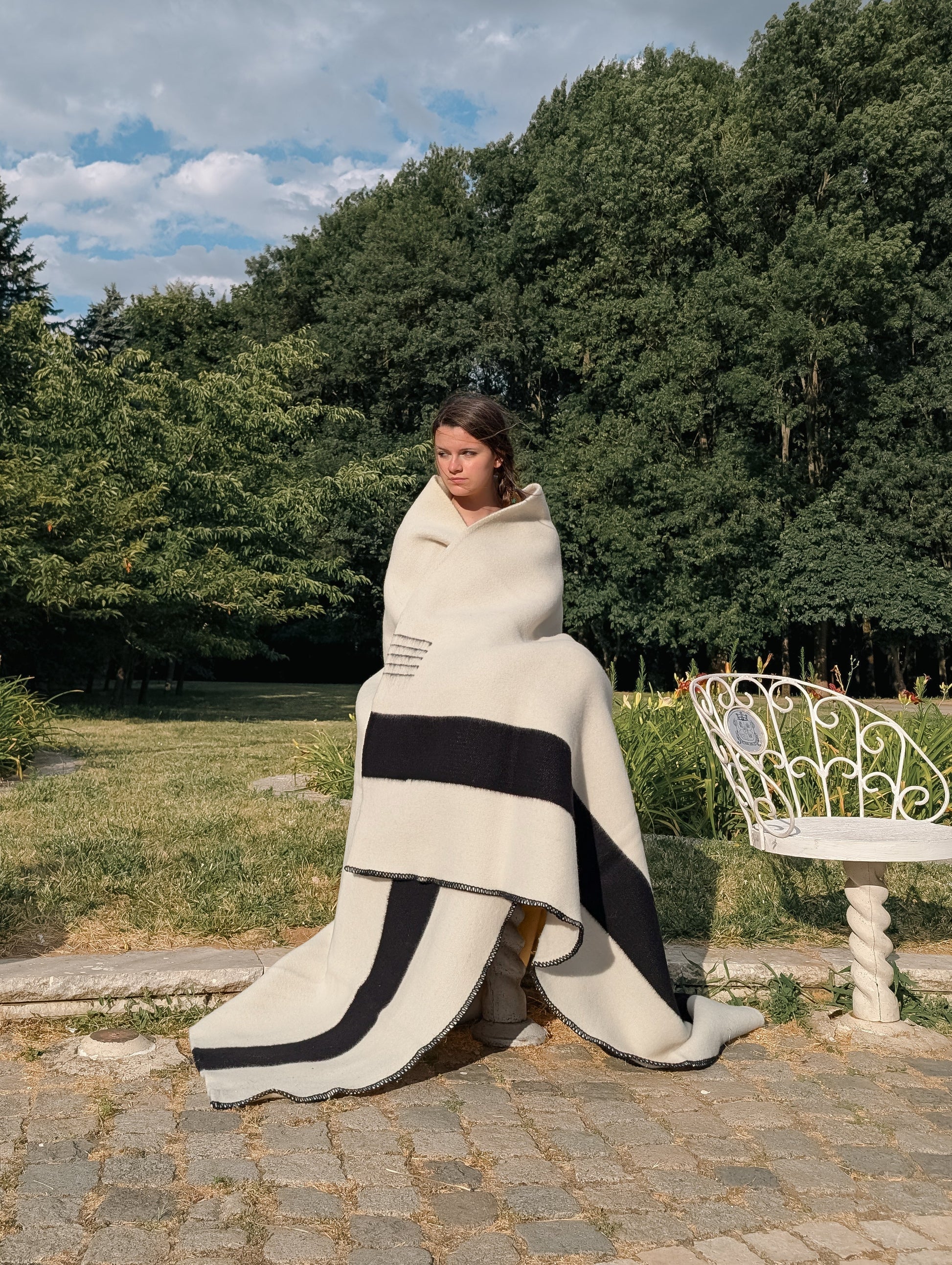 Girl sitting covered with a heirloom cream blanket with black stripes in an outdoors settinng with nature 