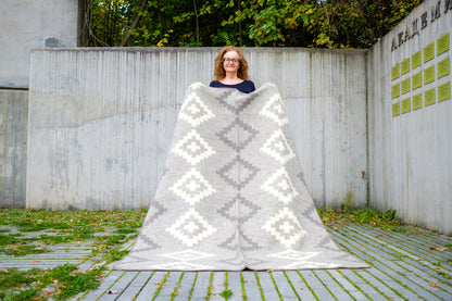 Woman standing outdoors with a large undyed gray and white patterned blanket draped over her. The blanket is handmade from premium quality new pure wool.