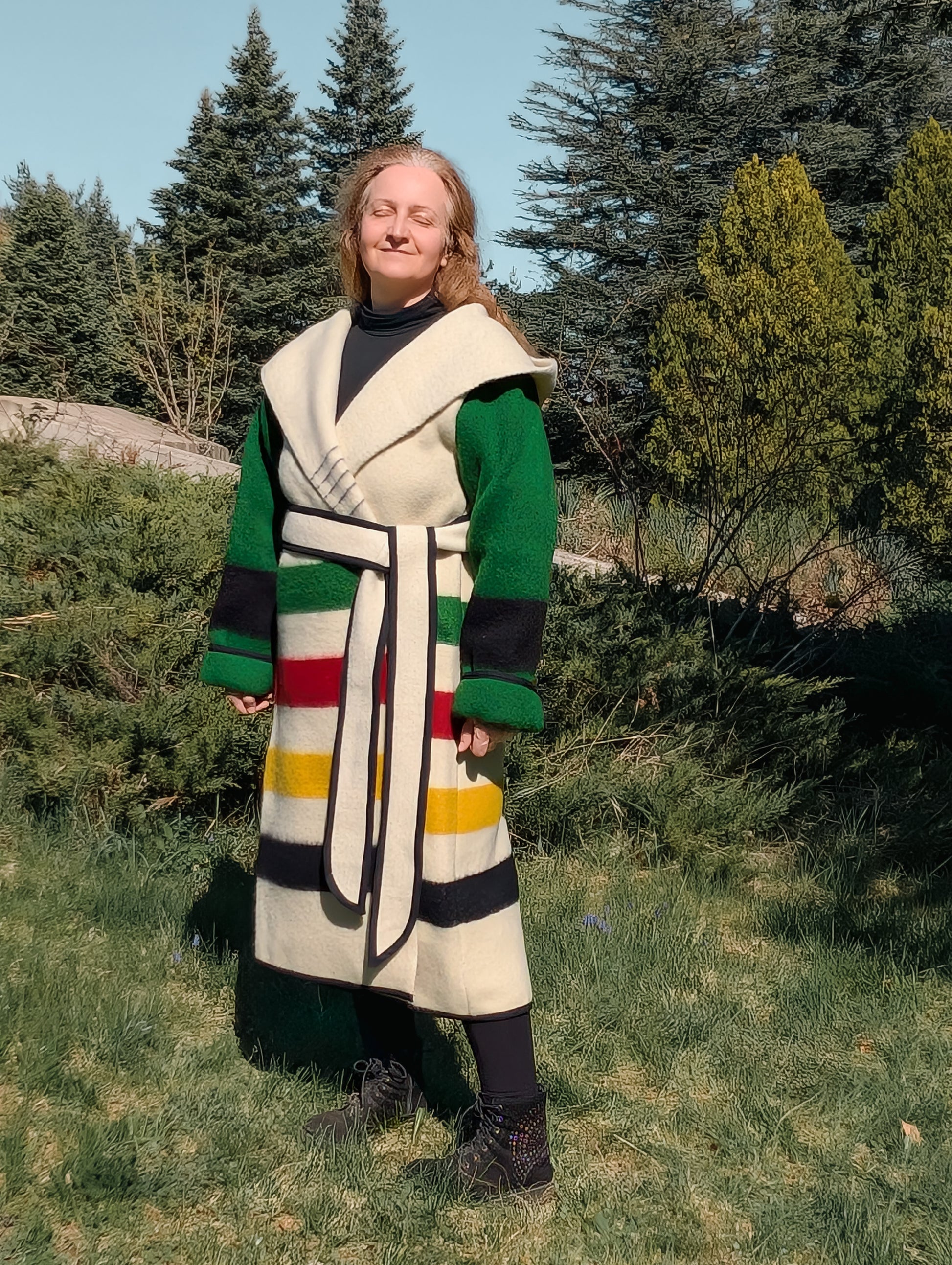 A woman posing with a wrap capote coat with a large hood, cinched with a belt. Made from repurposed stripe wool blanket.