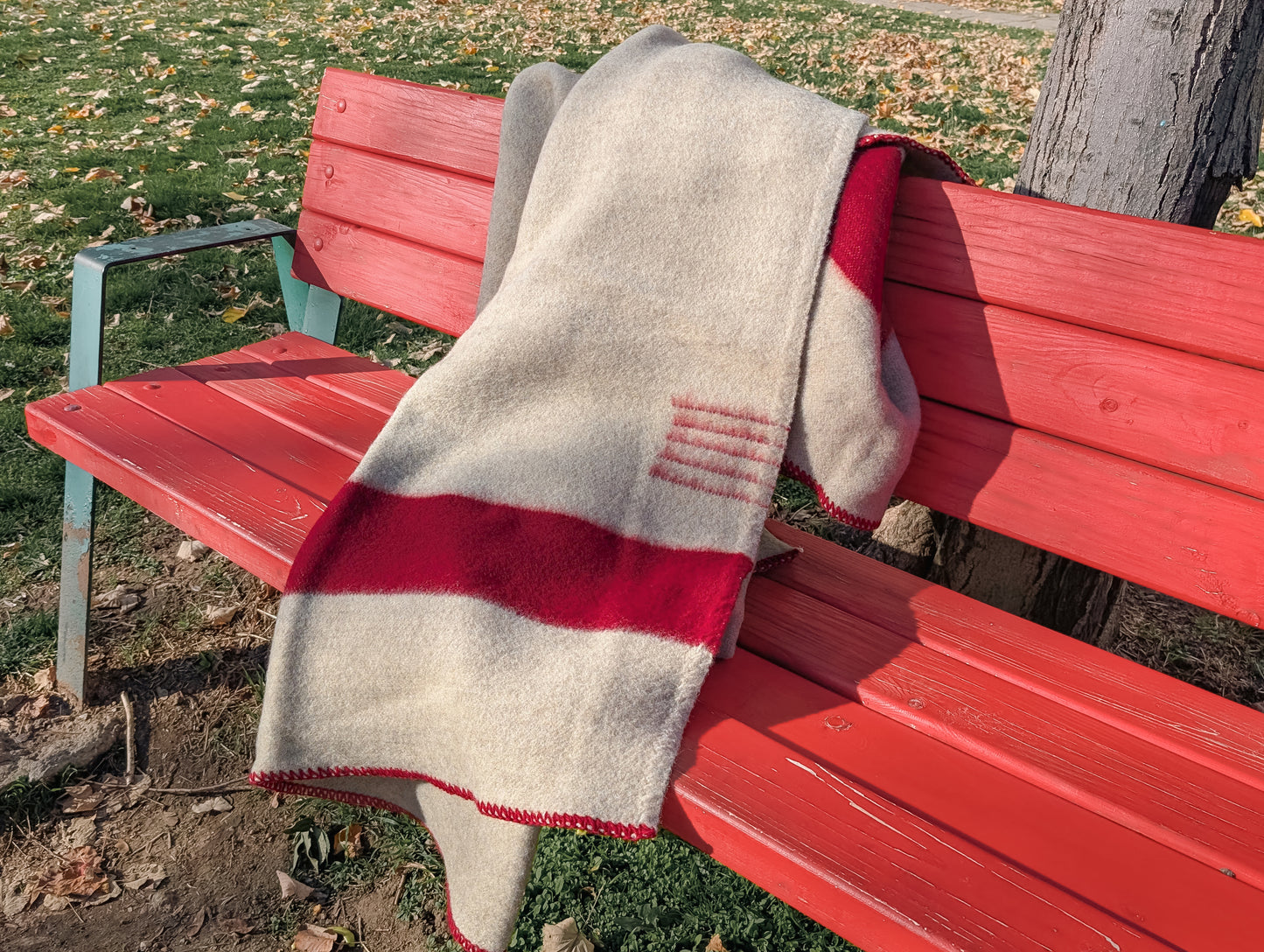 Premium organic gray wool blanket with red stripes, draped on a red bench outside in a park.