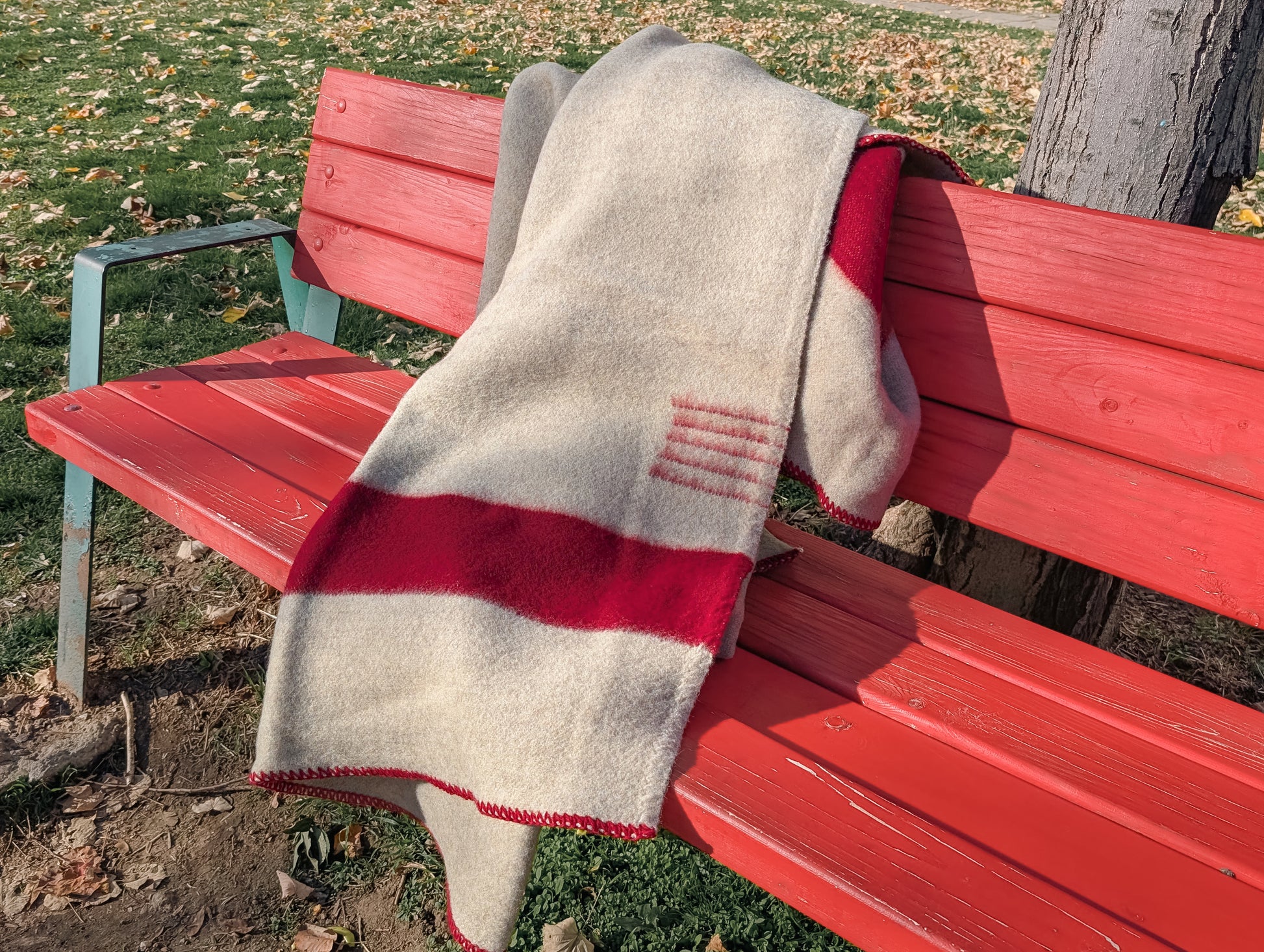 Premium organic gray wool blanket with red stripes, draped on a red bench outside in a park.