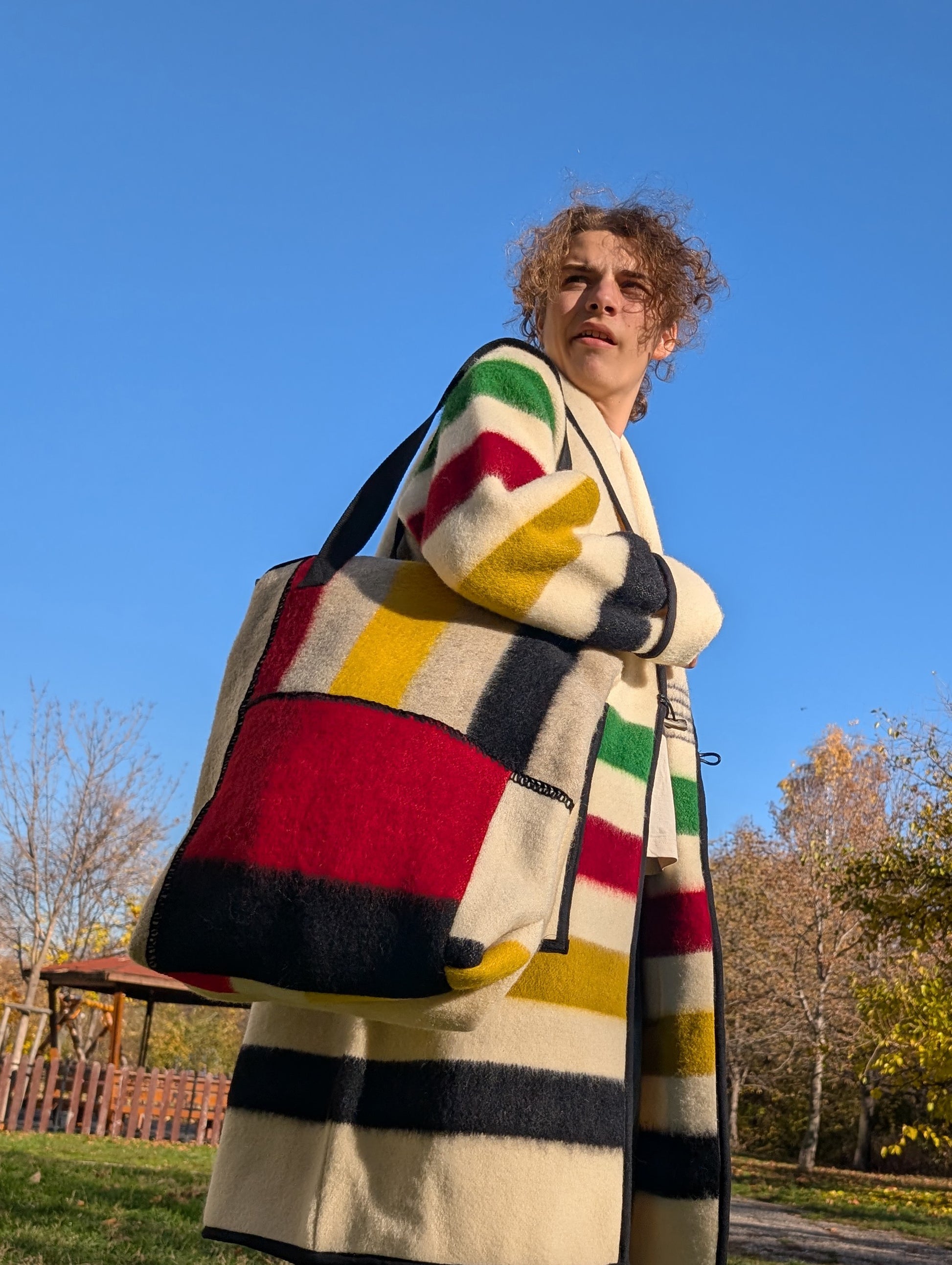 Young man wearinga a colorful patterned unisex coat and a matching unisex large shoulder bag made of wool blanket leftovers. 