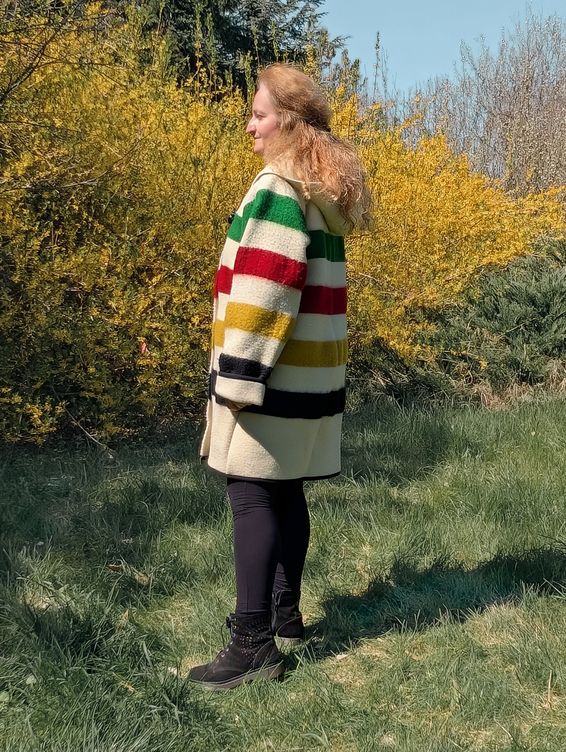 A woman posing outside with greenery behind. The woman is wearing a midi swing wool blanket coat witha shawl collar hoodie and toggle closure. 