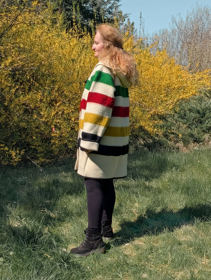 A woman posing outside with greenery behind. The woman is wearing a midi swing wool blanket coat witha shawl collar hoodie and toggle closure. 
