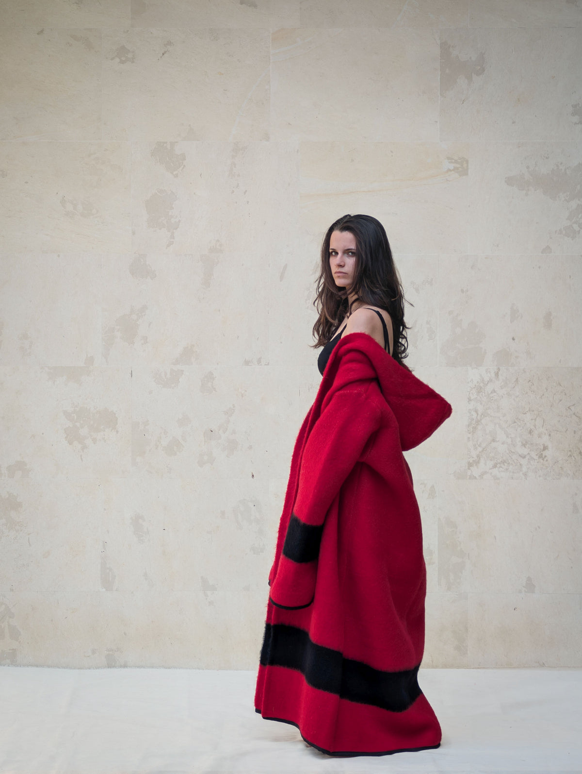 Side view of long red handwoven wool coat with hood, black stripe detail and fluid oversized silhouette