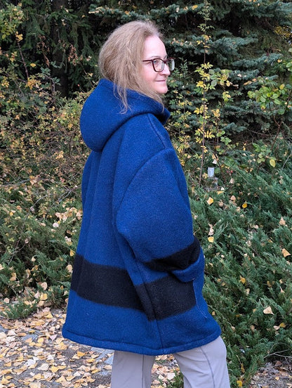 A woman dressed with blue striped Boreal shirt, wool anorak, upcycled from a new pure wool blanket, with a wide hood and a big front kangaroo pocket. Oversized unisex model.
