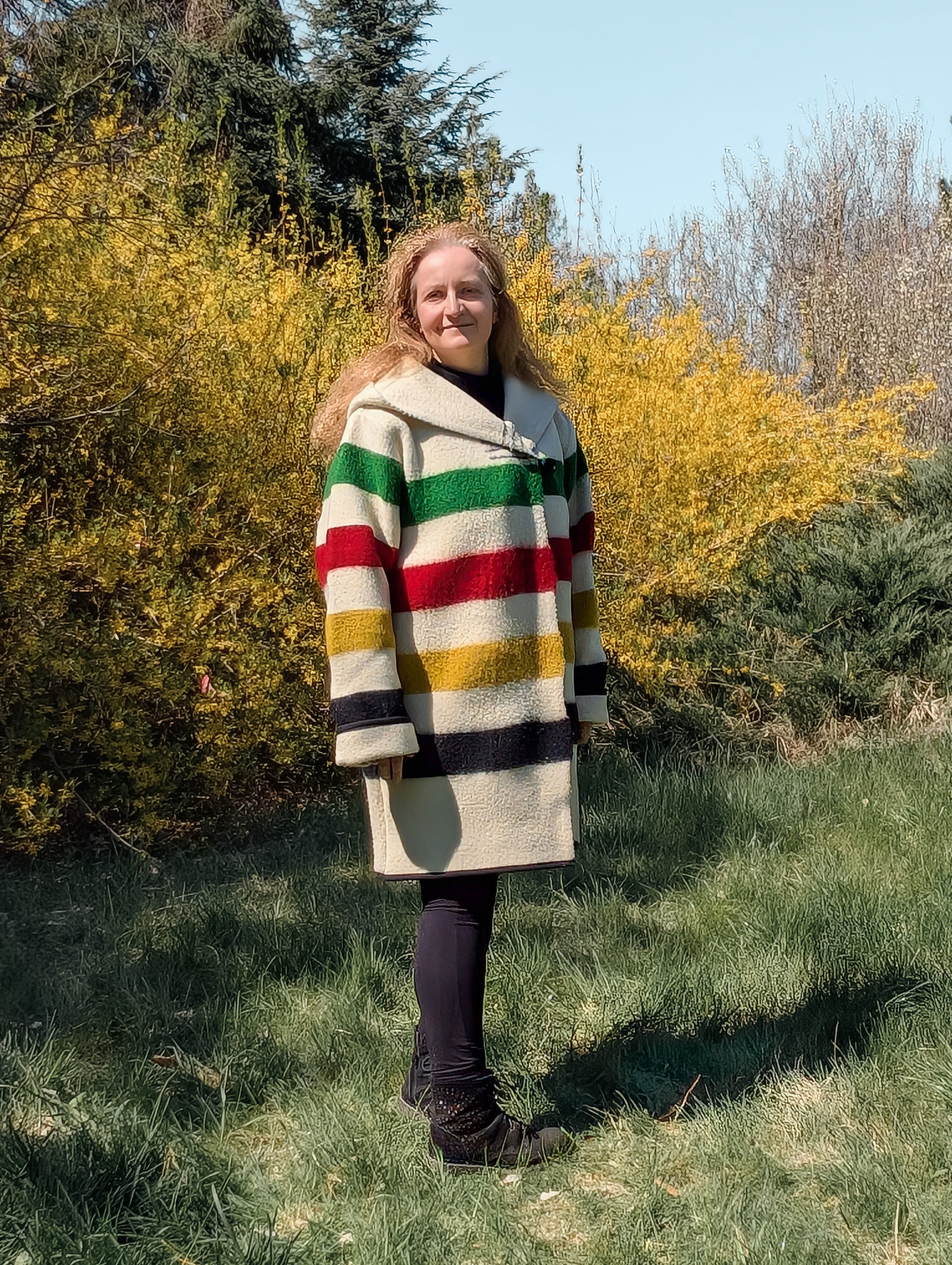 Person wearing a colorful striped coat standing in a grassy area with trees and bushes. The person is wearing a loose comfortable midi size wool hooded coat. 