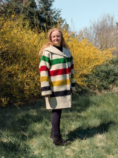 Person wearing a colorful striped coat standing in a grassy area with trees and bushes. The person is wearing a loose comfortable midi size wool hooded coat. 