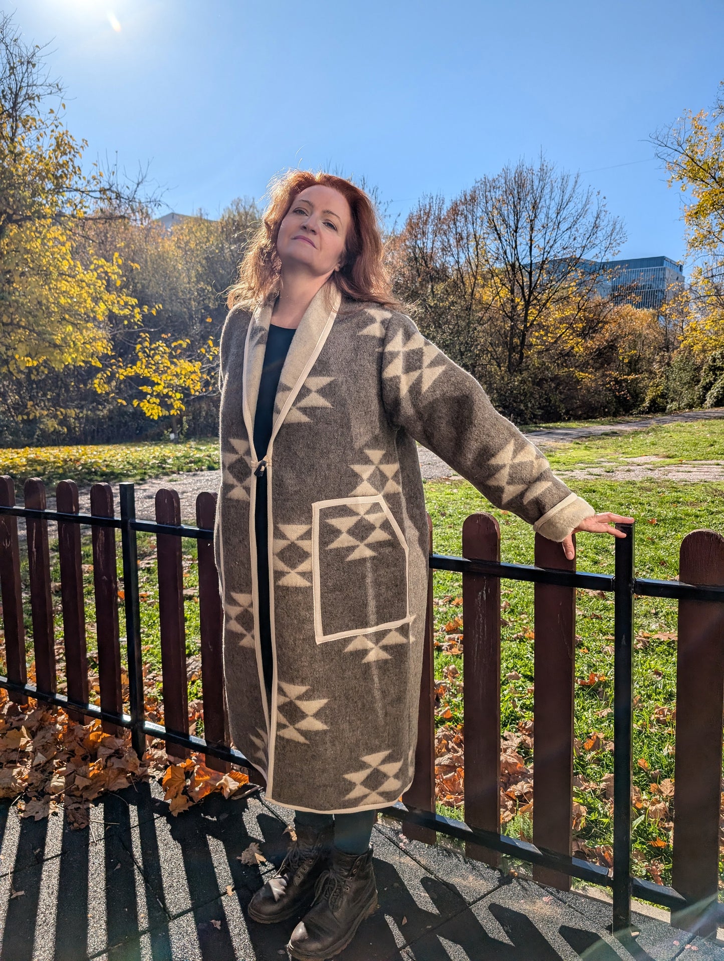 A woman leaning on the fence is wearing a tailored long wool cardigan coat made from undyed handmade wool blanket with folk geometric motifs.