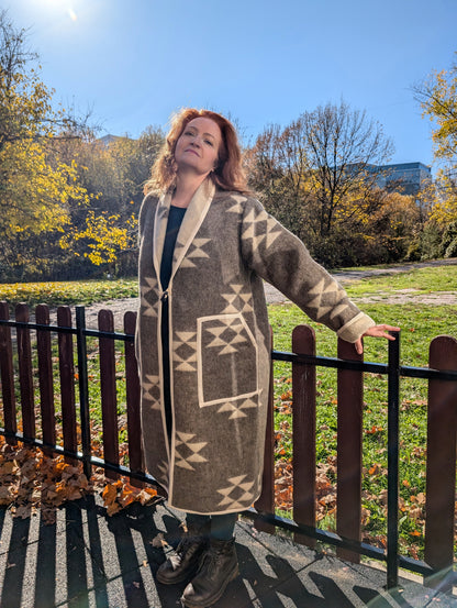A woman leaning on the fence is wearing a tailored long wool cardigan coat made from undyed handmade wool blanket with folk geometric motifs.