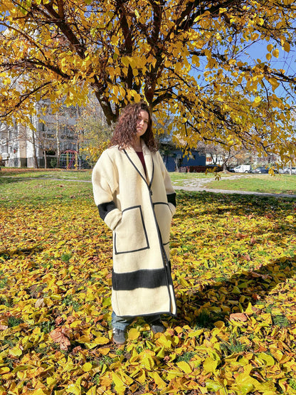 A woman wearing a loose ivory kimono coat upcycled from a natural virgin woolen blanket with black stripes. The coat features a cardigan collar, two front pockets and a toggle button. 