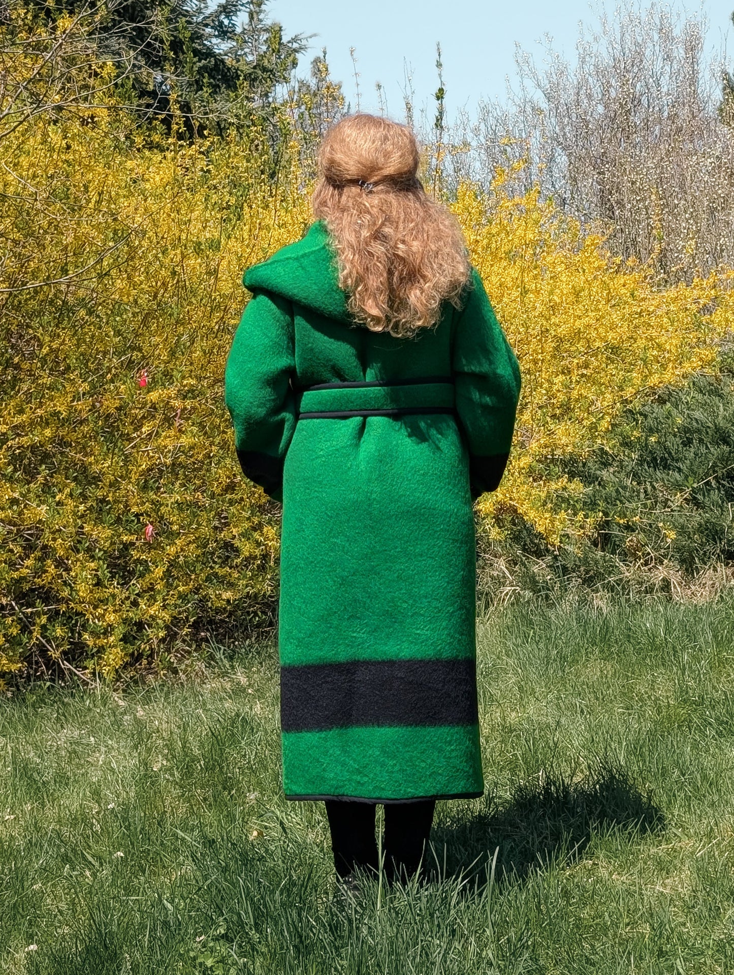Model posing backwards with a warm heritage slow fashion capote coat in green, with a black stripe, wrapped with a belt.
