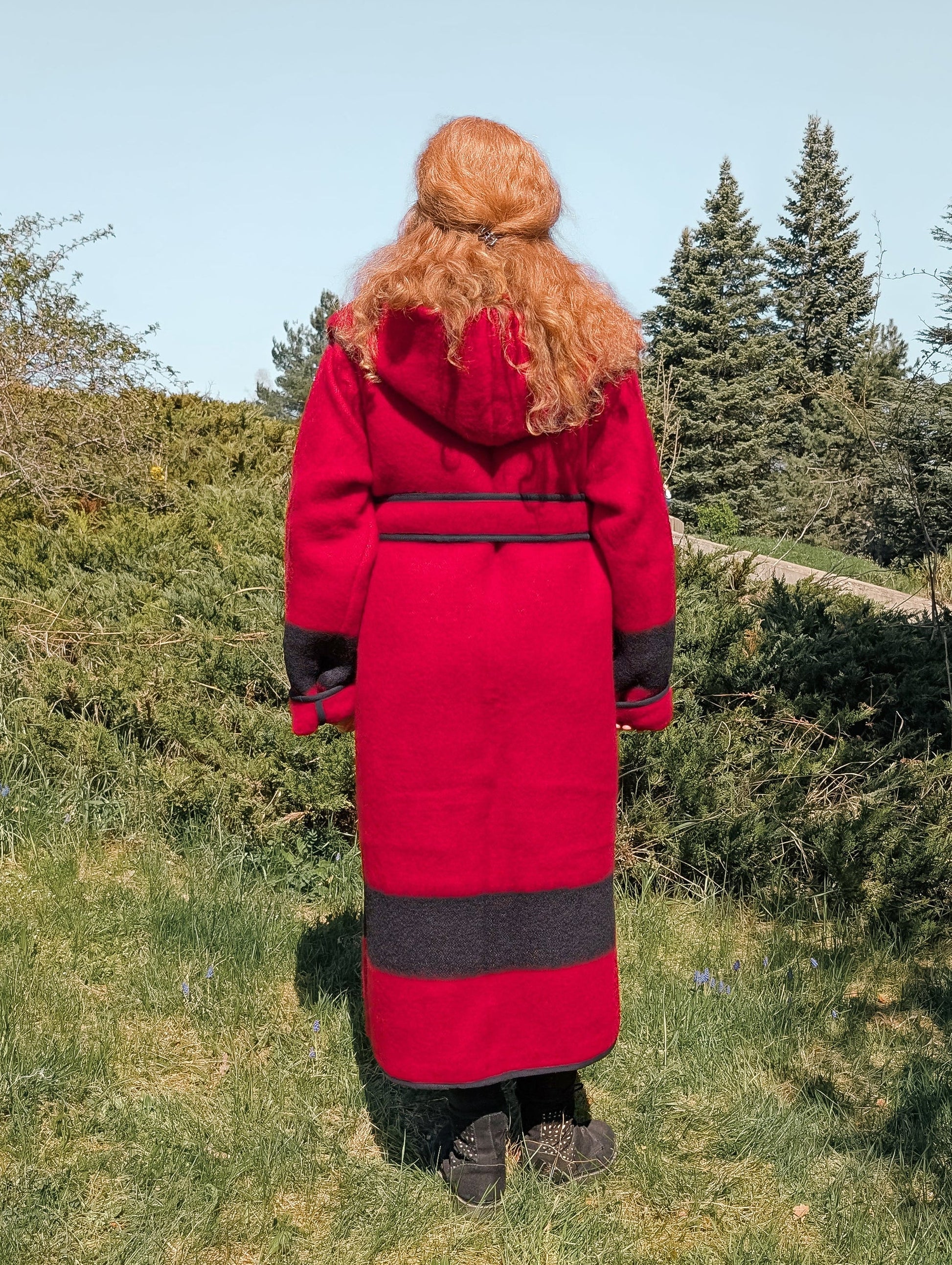 A woman posing backwards with a custom made long wool jacket in red with black stripes, hood and belt.